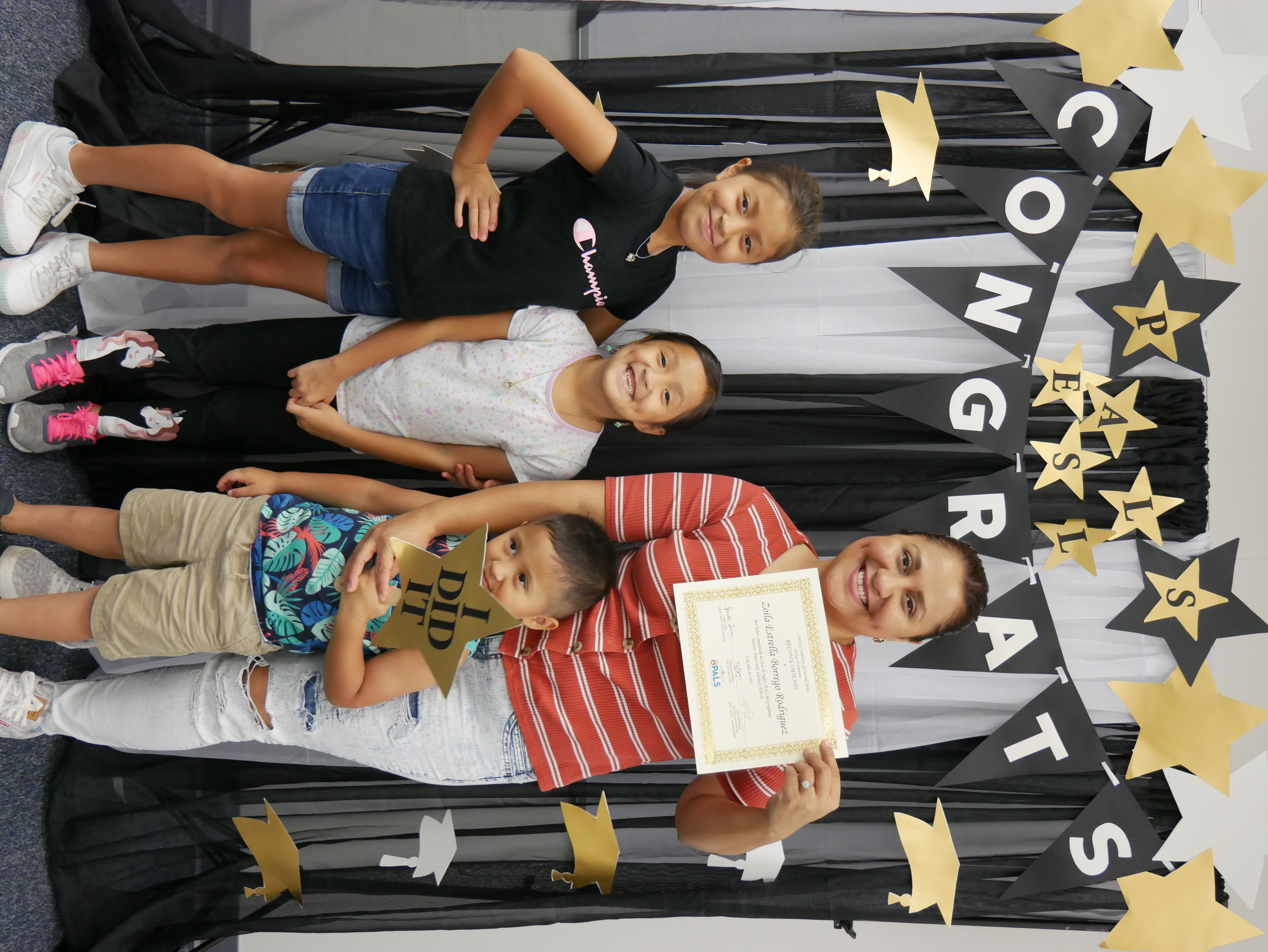 woman standing with her children (daughter 9, daughter 7, son 3), she is smiling and holding her english language class graduation certificate in celebration of her accomplishment
